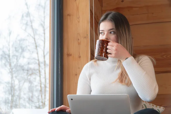 A Woman Sipping Coffee And Working On Her Laptop In A Cozy Room - Sitting Next To Window