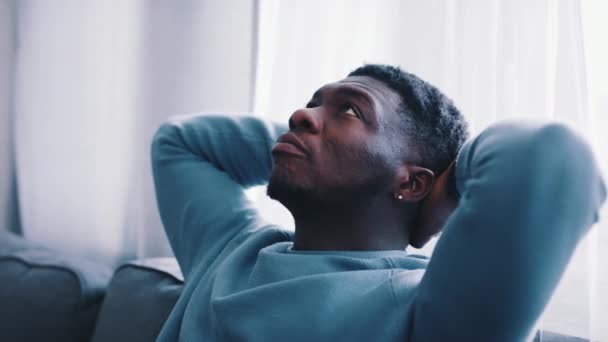 Hombre serio pensando mientras está sentado en un sofá azul - Las manos detrás de la cabeza — Vídeos de Stock