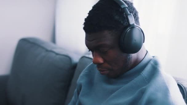 Homem afro-americano focado usando fones de ouvido sentado em um sofá — Vídeo de Stock