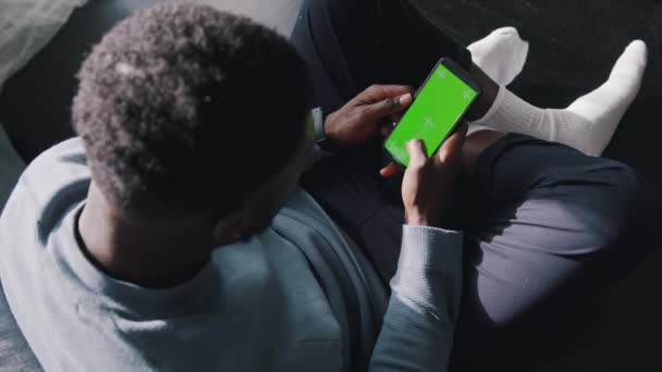 Hombre sentado con las piernas cruzadas en el sofá usando un teléfono celular con una pantalla verde. — Vídeos de Stock