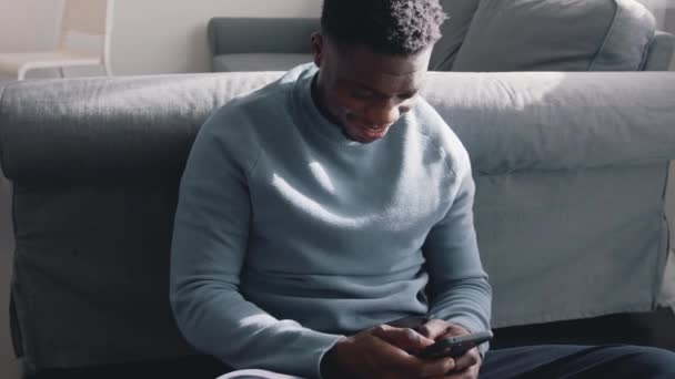 Hombre negro afroamericano sentado en el suelo en una acogedora habitación en casa usando su teléfono celular — Vídeo de stock