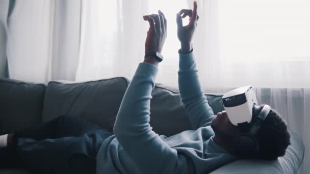 Αφρο-Αμερικανός άνδρας με γυαλιά εικονικής πραγματικότητας και ακουστικά ξαπλωμένα στον καναπέ — Αρχείο Βίντεο