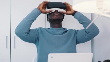 Siyah bir adam sanal gerçeklik gözlüğü takıyor ve laptopuyla masada oturuyor.