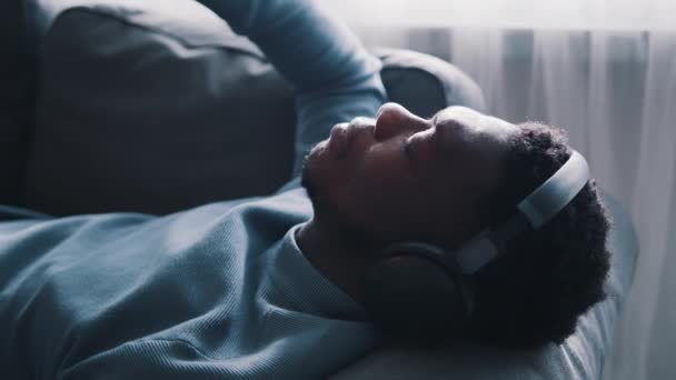 Homem afro-americano a usar auscultadores a ouvir música. Deitado no sofá — Vídeo de Stock