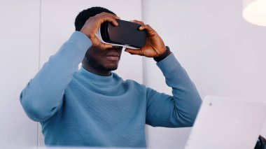 Sanal gerçeklik gözlüğü takan siyahi bir adam laptopuyla masada oturuyor.