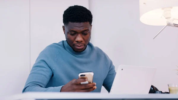 Hombre negro afroamericano con expresión de cara seria usando el teléfono delante de la computadora portátil. — Foto de Stock