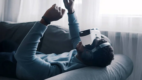 Homem afro-americano usando óculos de realidade virtual e fone de ouvido deitado no sofá. Um homem a trabalhar com óculos de realidade virtual a tocar em alguma coisa. Concentrado. — Fotografia de Stock