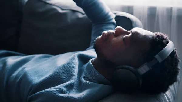 Αφροαμερικάνος που φοράει ακουστικά ακούγοντας μουσική. Ξαπλωμένος στον καναπέ. — Φωτογραφία Αρχείου