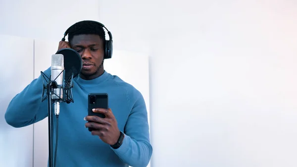 Kulaklık takan bir adam stüdyoda mikrofona şarkı söylüyor. Elinde bir telefon tutarak sözlere bakıyor.. — Stok fotoğraf