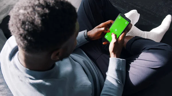 Homem sentado de pernas cruzadas no sofá usando um telefone celular com uma tela verde. — Fotografia de Stock