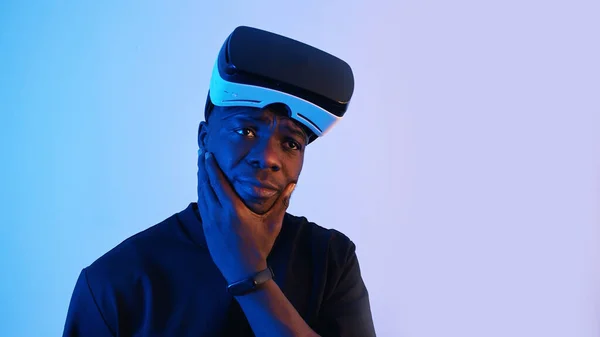 Афро-американец в очках виртуальной реальности на лбу, осматривается. Думаю кое о чем. Виртуальные наушники. Синий фон. Студия. — стоковое фото