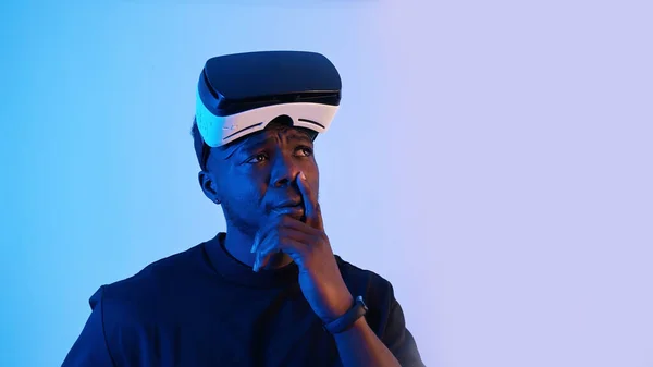 Афро-американец в очках виртуальной реальности на лбу, осматривается. Думаю кое о чем. Виртуальные наушники. Синий фон. Студия. — стоковое фото