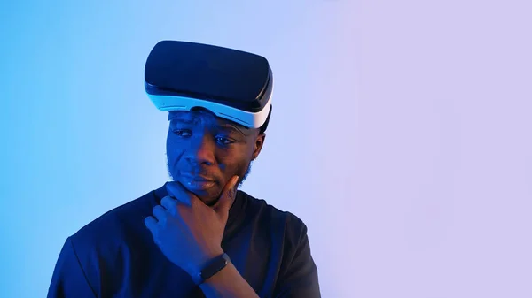 Um afro-americano com óculos de realidade virtual na testa a olhar à volta. Estou a pensar em alguma coisa. óculos de fone de ouvido virtual. Fundo azul. Imagens do estúdio. — Fotografia de Stock