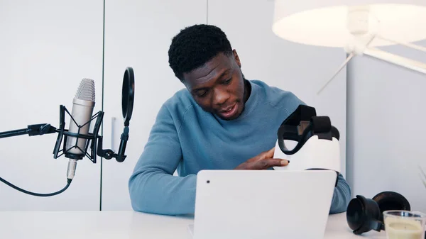 Hombre sosteniendo unas gafas VR sentado en la mesa con un portátil y un micrófono — Foto de Stock