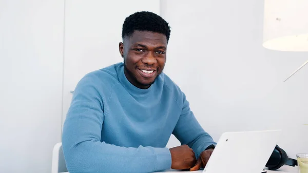 Hombre negro afroamericano sentado en la mesa frente a una computadora portátil trabajando y sonriendo a la cámara — Foto de Stock