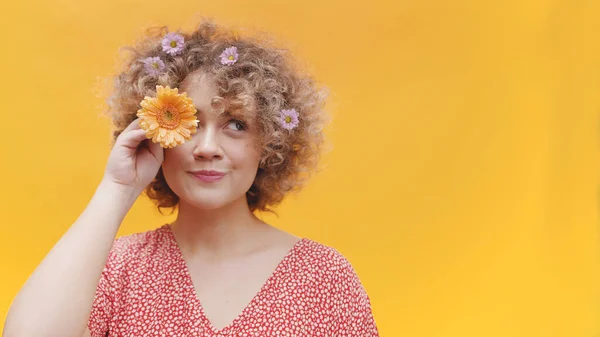 Playful young woman with curly hair holding flower over her eye — Stockfoto