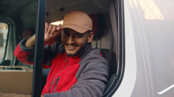Delivery guy wearing red uniform sitting in the white van and tipping his cap