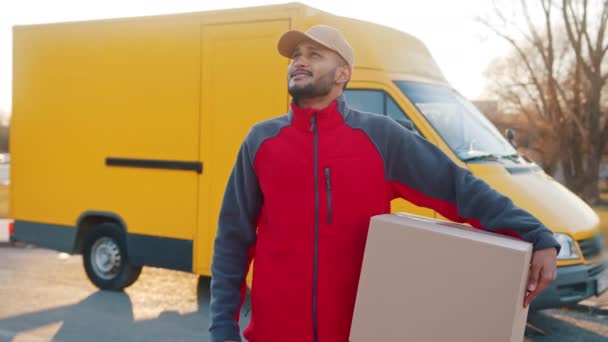 Rapaz de entrega carregando uma caixa de papelão um caminhão amarelo atrás dele - entrega do pacote — Vídeo de Stock