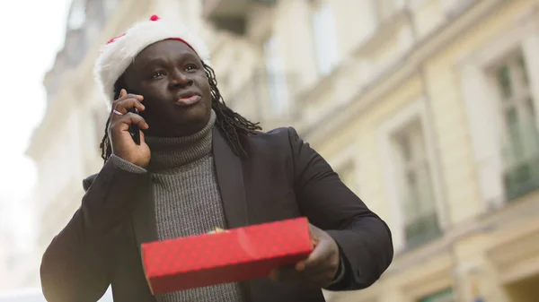 Siyah bir adam elinde paketlenmiş bir hediyeyle cep telefonunu arıyor. — Stok fotoğraf