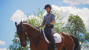 Genç bir kız, doğada bir ağacın yanında kahverengi bir atın üzerinde oturuyor.