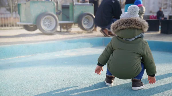 A small child with open fists squats in the middle of a playground near people — Stock Photo, Image