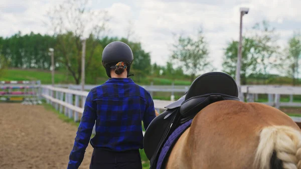 Uma jovem jóquei menina leva um cavalo ao lado de uma cerca de madeira para obstáculos — Fotografia de Stock