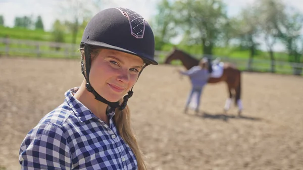 頭にヘルメットを被った少女が茶色の馬の横に立っているぼやけた女の子を見てカメラを見て — ストック写真