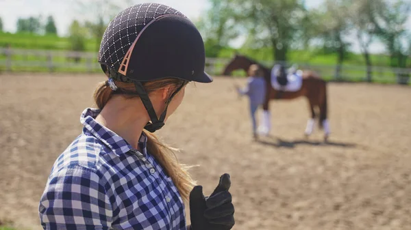 Uma jovem com um capacete na cabeça olha para uma garota borrada de pé ao lado de um cavalo marrom — Fotografia de Stock