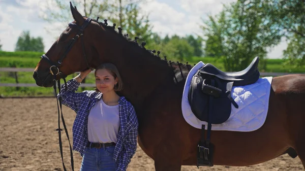 A young girl in an unbuttoned shirt in a white T-shirt stands under a horses neck and scratches his head — Stock Photo, Image