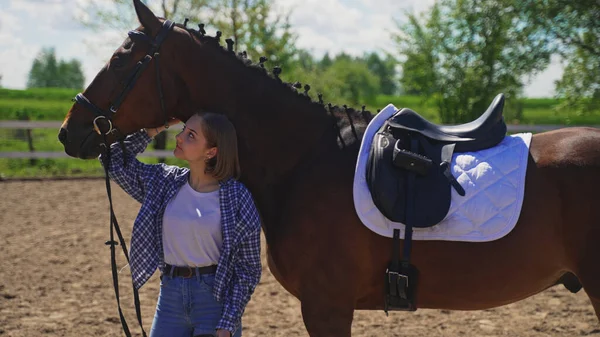 A young girl in an unbuttoned shirt in a white T-shirt stands under a horses neck and scratches his head and looks at him — Stock Photo, Image