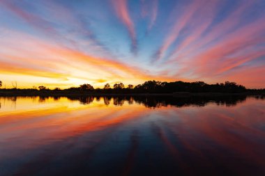 Nehrin üzerinde gün batımı, ormanın silueti, suda ayna görüntüsü.