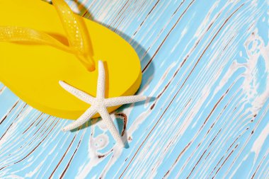 Sarı kauçuk plaj terlikleri ve mavi ahşap arka planda deniz yıldızı, metnin altına.