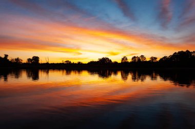 Nehrin üzerinde gün batımı, ormanın silueti, suda ayna görüntüsü.