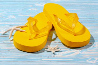 Sarı kauçuk plaj terlikleri ve mavi ahşap arka planda deniz yıldızı, metnin altına.