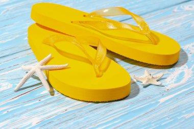 Sarı kauçuk plaj terlikleri ve mavi ahşap arka planda deniz yıldızı, metnin altına.