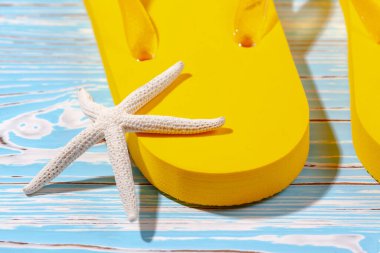 Sarı kauçuk plaj terlikleri ve mavi ahşap arka planda deniz yıldızı, metnin altına.