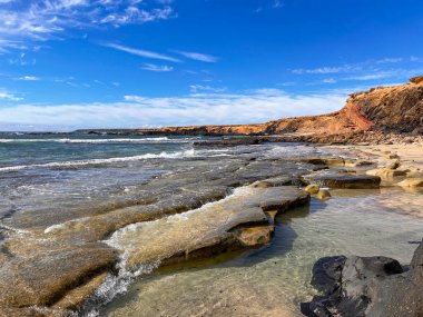 Playa de Los Ojos 'un yüksek çözünürlüklü fotoğrafı, Fuerteventura, Kanarya Adaları' nın güneybatı kıyısında. Görüntü kayalık oluşumları, karanlık volkanik uçurumları ve doğal gün ışığı altında kum, kaya ve denizin zıt tonlarını vurgular..