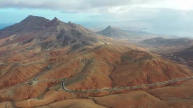 Sicasumbre dağlarının ve Fuerteventura 'daki dolambaçlı yolun hava aracı görüntüsü, açık mavi gökyüzünün altındaki volkanik manzaraları ve çöl tepelerini gösteriyor..