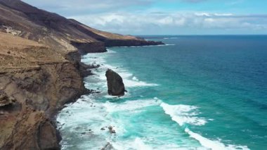 Fuerteventura, Kanarya Adaları 'ndaki Coffee Beach' in sersemletici hava görüntülerinde karamsar, bulutlu bir gökyüzü altında ikonik Roque del Moro kaya oluşumu görülüyor. Uzak ve dramatik bir Atlantik manzarası.