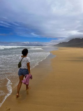 Fuerteventura, Kanarya Adaları 'ndaki vahşi Coffee Plajı' nda gezen genç bir kadın. Güneş ve bulutların altında küçük bir çapraz vücut çantası taşıyor..