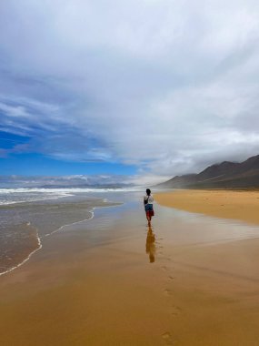 Fuerteventura, Kanarya Adaları 'ndaki vahşi Coffee Plajı' nda gezen genç bir kadın. Güneş ve bulutların altında küçük bir çapraz vücut çantası taşıyor..
