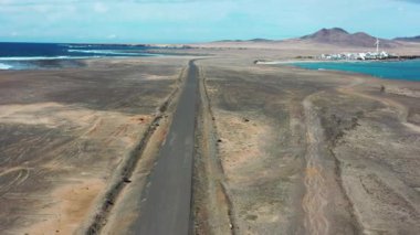 Punta de Jandia 'ya giden uzak yolu ve güney Fuerteventura' nın engebeli manzarasına karşı kurulan El Puertito balıkçı köyünü kapsayan geniş bir drone perspektifi..