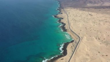 Güneşli bir günde, Corralejo Çölü 'nün yanındaki El Rosadero Sahili' nden geçen düz bir otoyolun hava görüntüsü. Hareket eden arabalar, altın kum tepeleri ve Atlantik kıyıları Fuerteventura 'daki bu insansız hava aracı videosunda çarpıcı bir kontrast oluşturuyor.