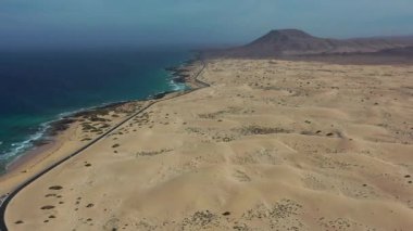 İspanya 'nın Kanarya Adaları, Fuerteventura' daki Corralejo çöl kumullarını geçen bir yolun insansız hava aracı görüntüleri. Manzara kurak arazi, kumlu dokular ve arka planda görünen ikonik Kızıl Dağ 'a sahiptir..