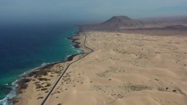 İspanya 'nın Kanarya Adaları, Fuerteventura' daki Corralejo çöl kumullarını geçen bir yolun insansız hava aracı görüntüleri. Manzara kurak arazi, kumlu dokular ve arka planda görünen ikonik Kızıl Dağ 'a sahiptir..
