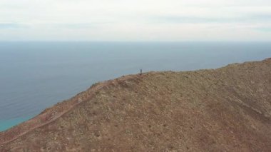 Fuerteventura, Kanarya Adaları 'ndaki Montaa Roja tepesinde duran bir çiftin insansız hava aracı görüntüleri. Volkanik kırmızı dağ, dramatik manzara, macera yolculuğu, özgürlük ve açık hava yaşam tarzı konsepti.