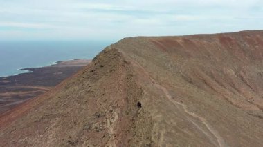 Sinema insansız hava aracı Fuerteventura 'daki Kızıl Dağ' ın dar sırtı boyunca inen bir kadın yürüyüşçüyü görüntülüyor. Engebeli volkanik oluşumlar ve geniş çöl dağlarıyla çevrili sahne yalnızlığı ve dayanıklılığı vurguluyor.,