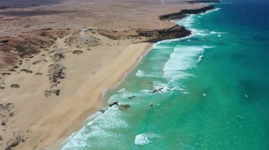 Yüksek açılı insansız hava aracı Playa del Castillo, Fuerteventura 'da uçurtma ve sörf aktivitelerini yakalıyor. Açık mavi gökyüzü, rüzgarla dolu uçurtmalar ve dalgalı okyanus dalgaları canlı bir yaz kıyı atmosferi yaratır..