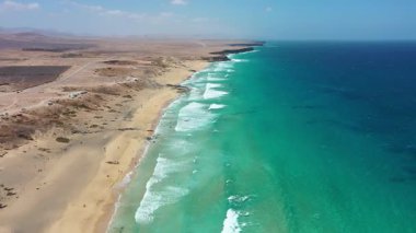 Uçurtmacılar ve sörfçülerin güneşli bir günde Atlantik Okyanusu 'nun tadını çıkardıkları Fuerteventura, Kanarya Adaları' ndaki Playa del Castillo 'nun insansız hava aracı görüntüsü. Altın kumlu plaj, turkuaz su ve aktif su sporları sahnesi.