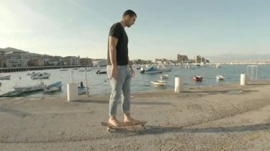 Kot pantolon ve kısa kollu siyah tişört giyen genç adam arka planda deniz olan iskelede yalın ayak kayak yapıyor.